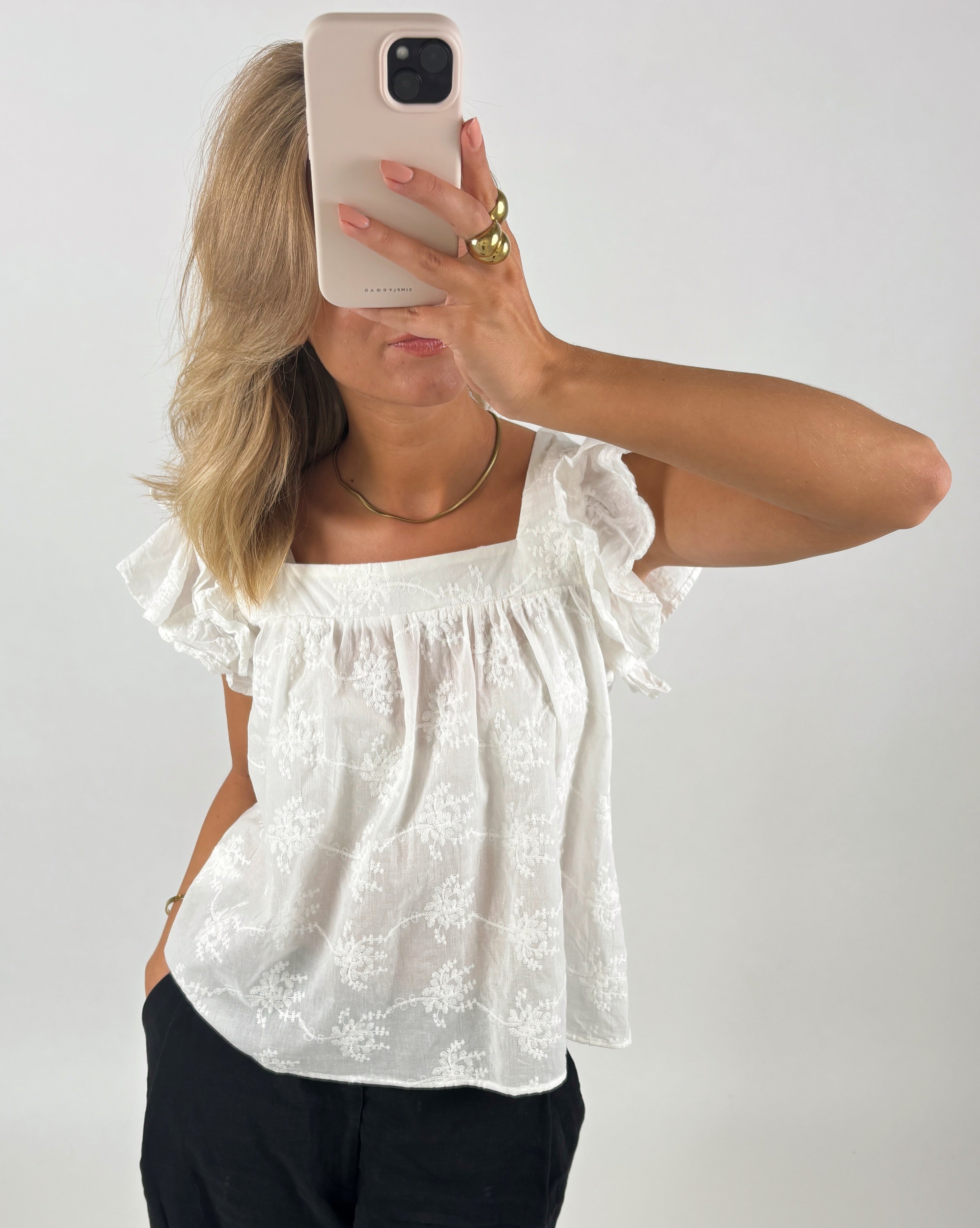 En person iført hvid, mønstret bluse tager et spejlselfie med en smartphone. Personen har let bølget, blond hår og guldtilbehør. Baggrunden er en ensfarvet væg.