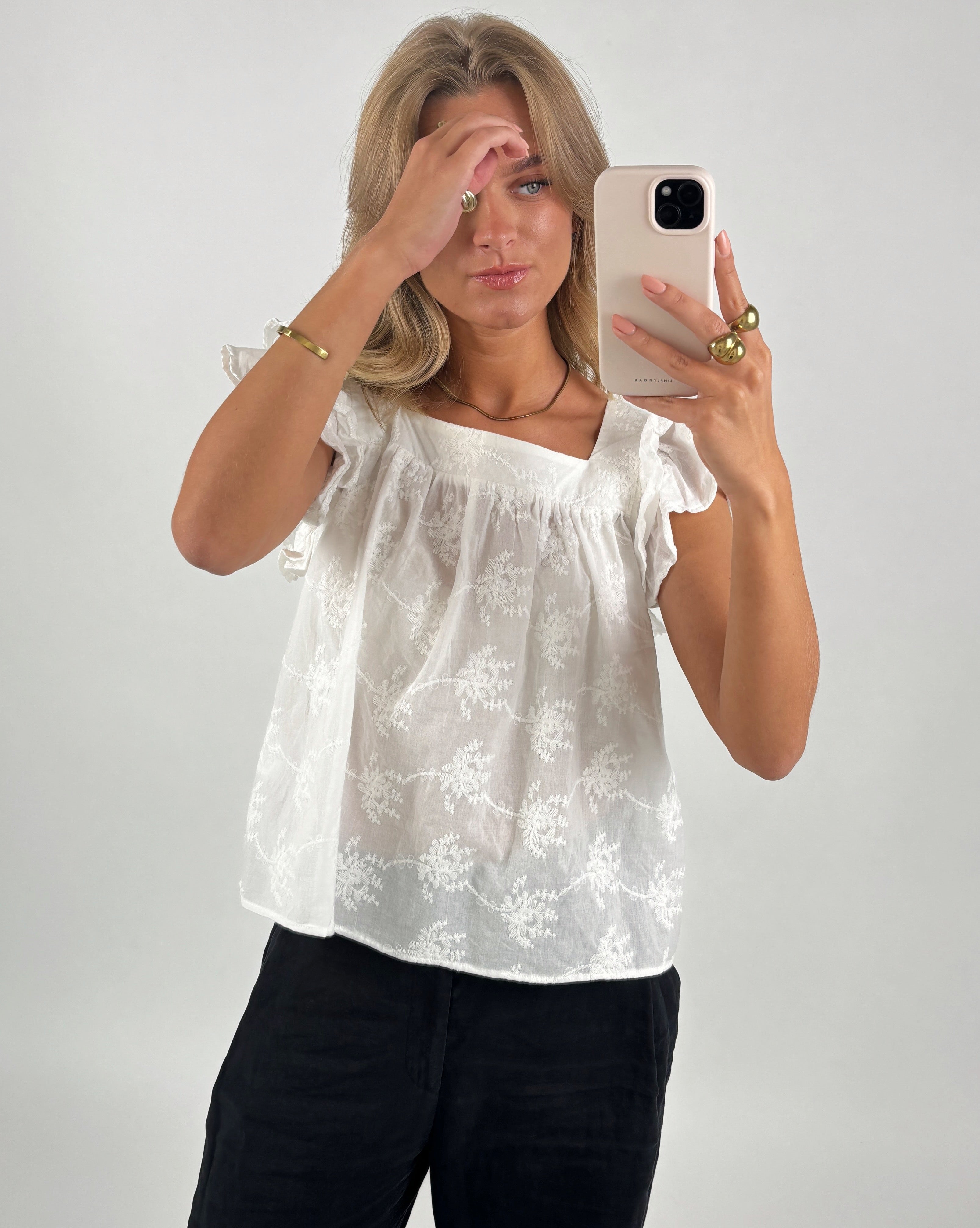 En person i en hvid bluse tager et spejlbillede med en smartphone. Personen rører ved panden med den ene hånd. Baggrunden er enkel og lys.