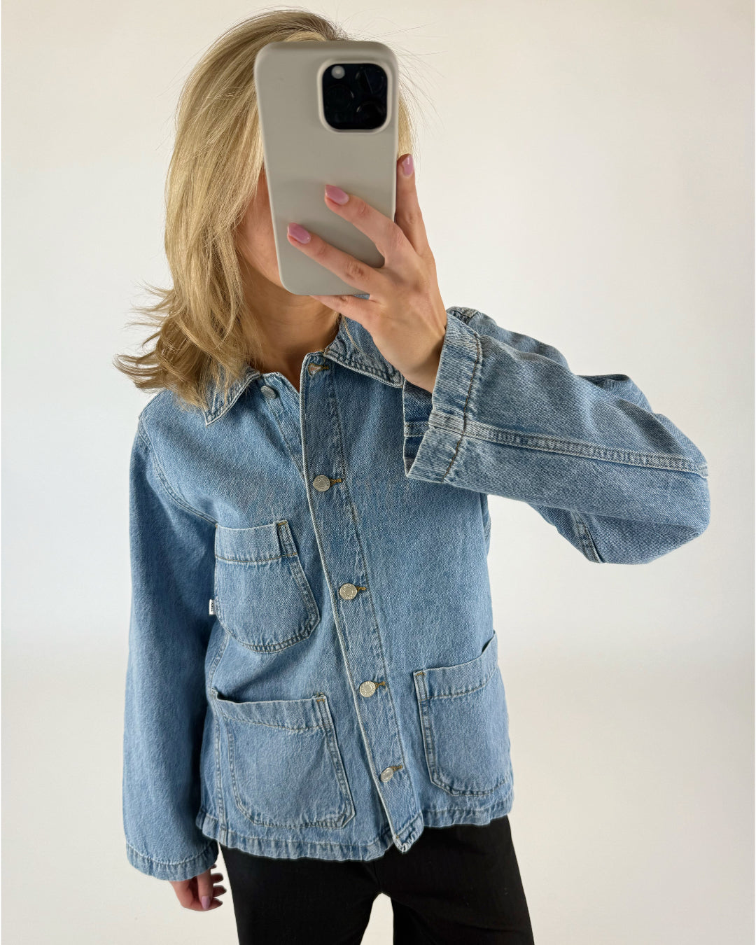 En person iført cowboyjakke tager en selfie i en minimalistisk, lys baggrund. Personen holder en smartphone foran ansigtet og har lyst hår og lyserøde negle.
