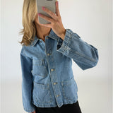 En person iført en blå denimjakke tager en selfie med en smartphone i et neutralt belyst rum. Jakken har store frontlommer og metalknapper.