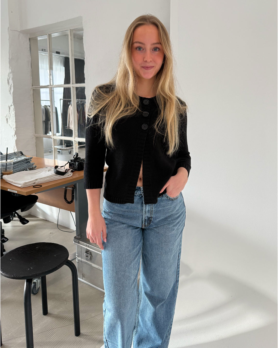 Kvinde står i en stue, iført sort bluse og blå jeans, ved siden af et bord med foldet tøj og et kamera. Væggene er hvide, og der er et stort vindue.