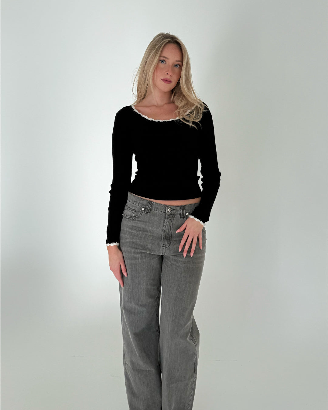 Kvinde står stille i et neutralt rum, iført sort trøje og grå jeans. Hun kigger fremad med afslappet kropsholdning. Der er ingen tekst i billedet.