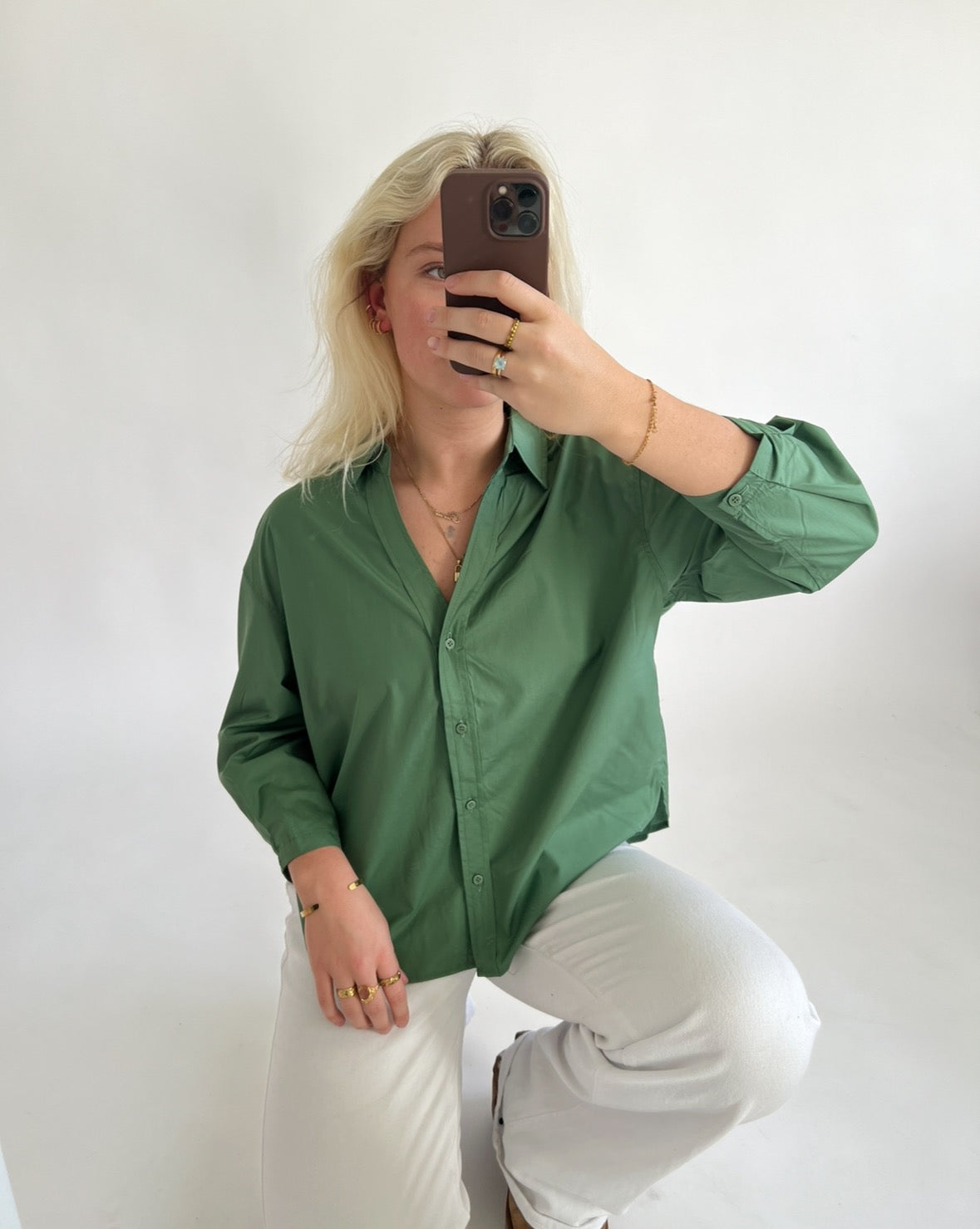 En person tager en selfie foran en hvid baggrund. Personen bærer en grøn skjorte og hvide bukser, med forskellige guldsmykker på fingrene og håndleddet.