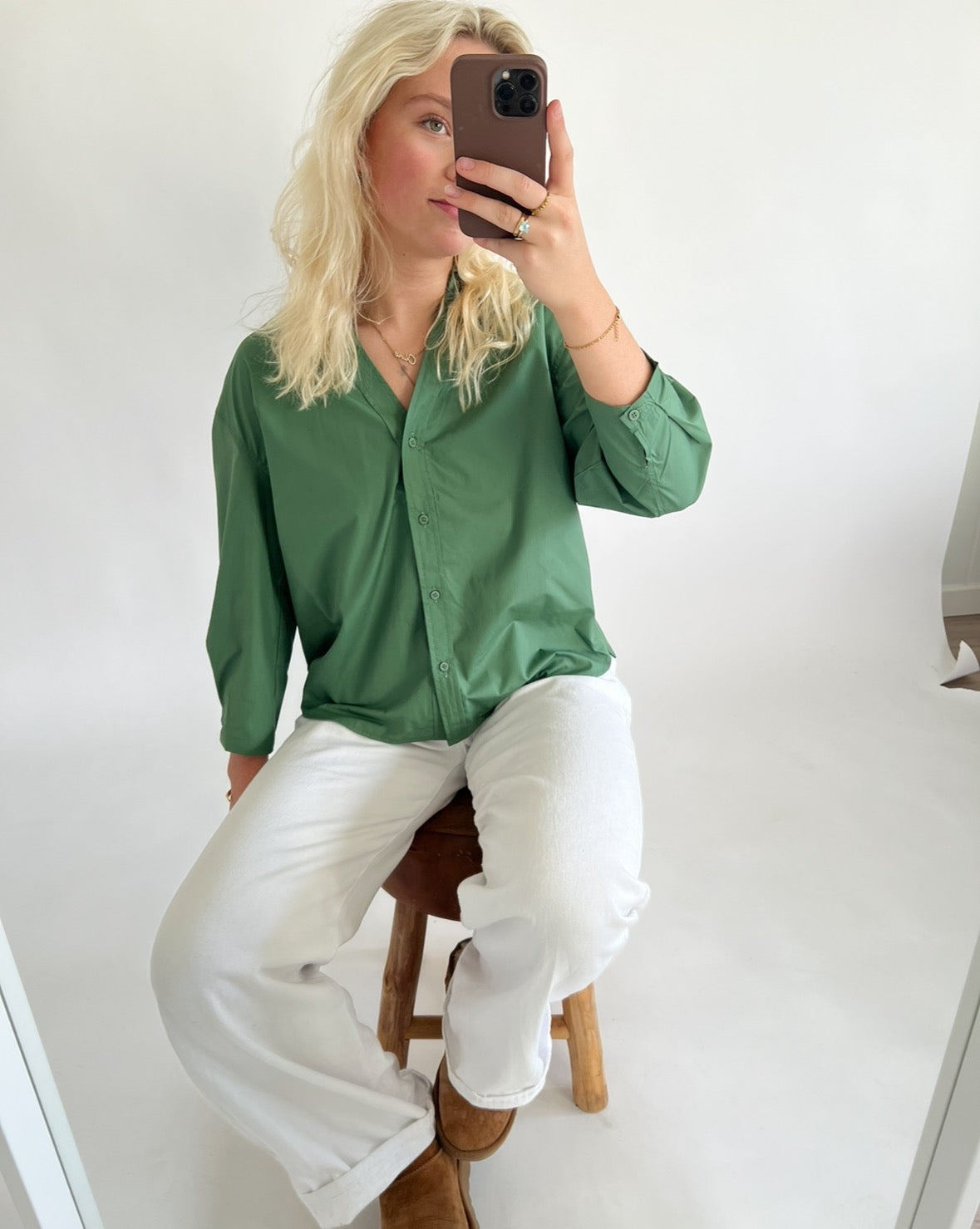 En person iført grøn skjorte og hvide bukser sidder på en træstol og tager en selfie i et minimalistisk rum med hvide vægge.