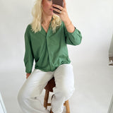 En person iført grøn skjorte og hvide bukser sidder på en træstol og tager en selfie i et minimalistisk rum med hvide vægge.