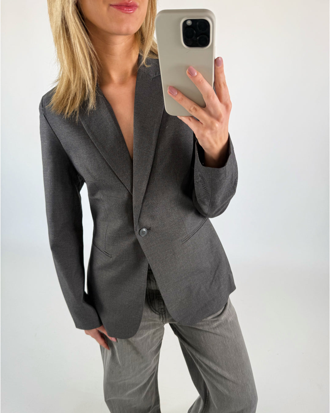 En person med blondt hår tager en selfie iført en grå blazer og bukser i lignende nuance. Baggrunden er en enkel, lys væg.