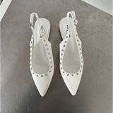 Hvide slingback-sko med spidse snuder dekoreret med sølvnitter, står på en metallisk overflade. Skoens indersål har teksten "stevemai PARIS".