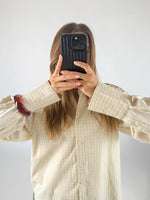 En person holder en mobiltelefon op foran ansigtet, iført en ternet skjorte og et rødt armbånd, i et lyst rum.