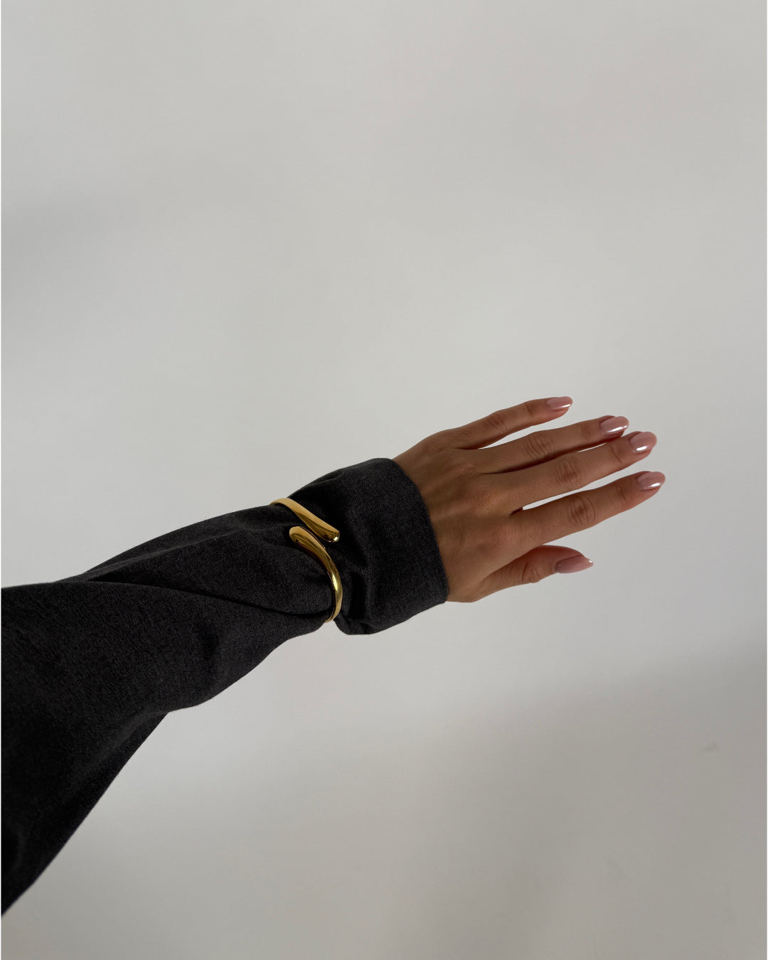En hånd med velplejede negle er let løftet, iført en guldfarvet armbånd, mod en neutral, lys baggrund.