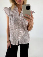 Kvinde iført blomstret skjorte med korte ærmer tager et spejlbillede med mobiltelefonen. Hun står i et hvidt rum og bærer sort bukser og et guldarmbånd.