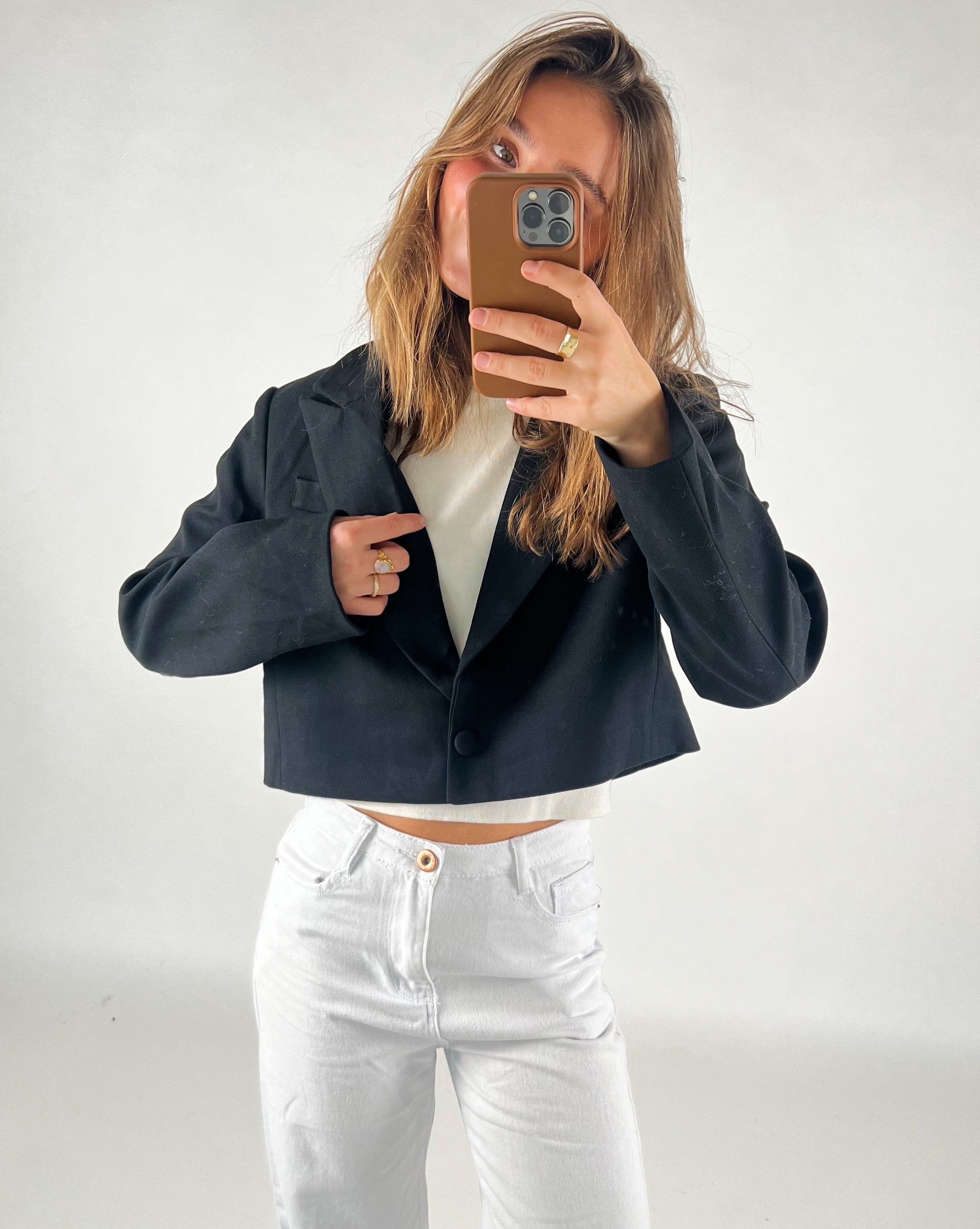 En person tager et spejlbillede iført en kort sort jakke og hvide bukser, mens der holdes en smartphone i et minimalistisk, lyst rum.