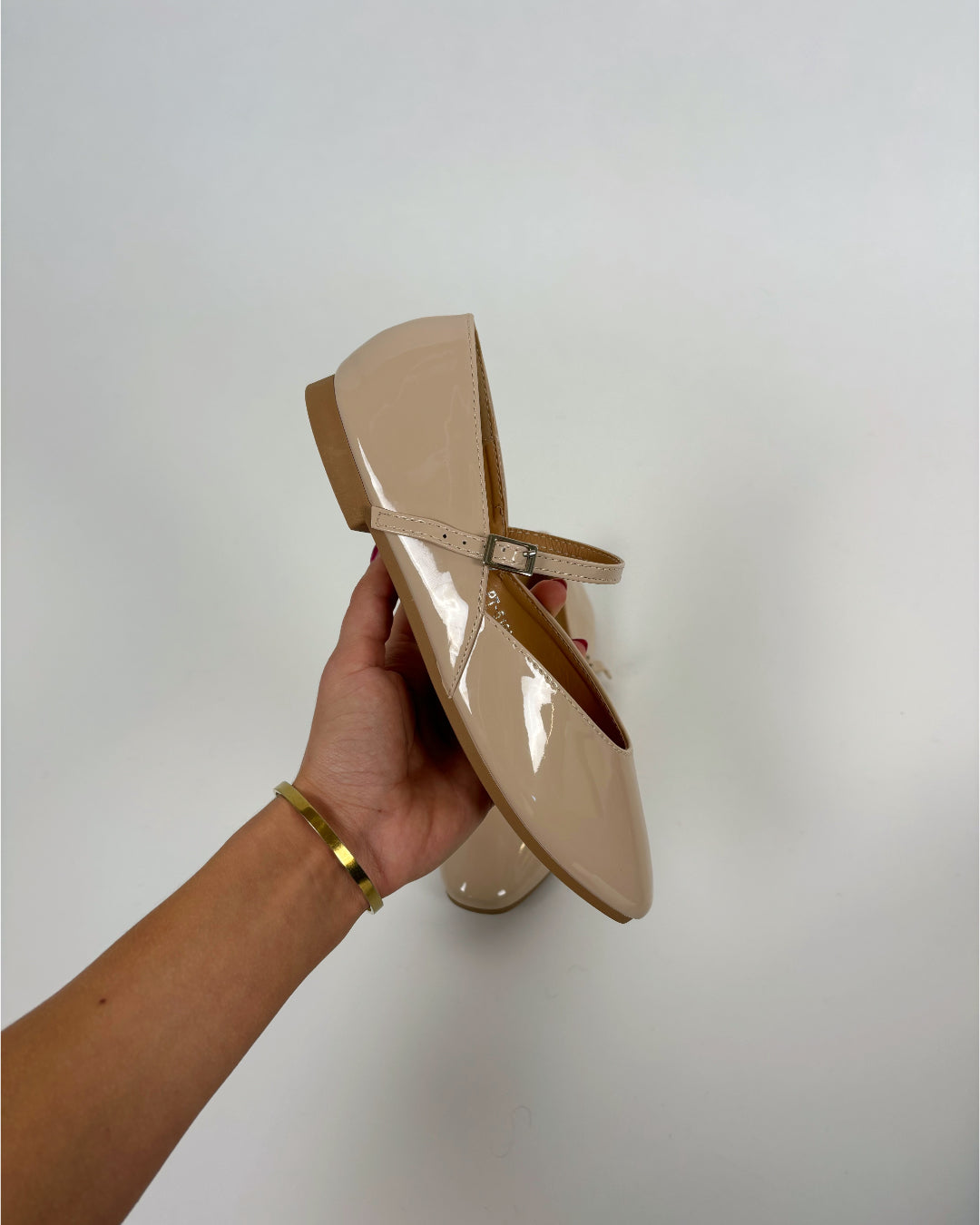 En hånd holder en beige lak-sko med rem og flad hæl mod en hvid baggrund. Personen bærer et guldfarvet armbånd som tilbehør.