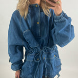 Person iført denimjakke tager selfie. Jakken har snører i taljen og lommer med trykknapper. Personen har lyst hår og bærer en gylden ring. Baggrunden er hvid.