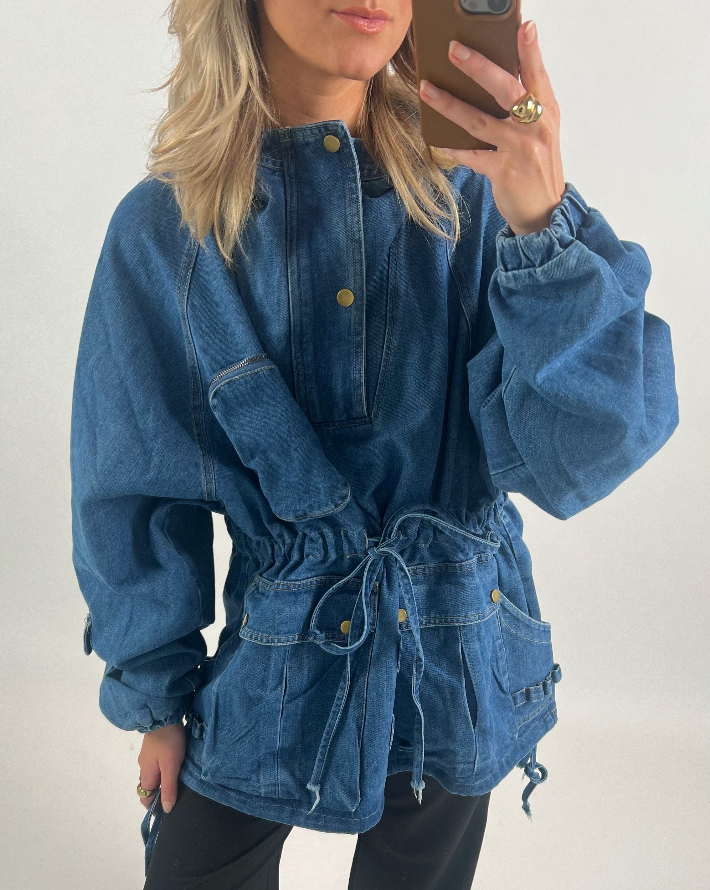 Person iført denimjakke tager selfie. Jakken har snører i taljen og lommer med trykknapper. Personen har lyst hår og bærer en gylden ring. Baggrunden er hvid.