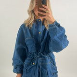 En person iført en stor, blå denimjakke med justerbare snore og lommer tager et spejlbillede med en mobiltelefon i et minimalistisk rum.