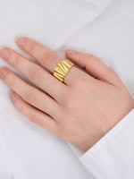 En hånd med en gylden ring med bølget design sidder afslappet på et hvidt klæde, delvist dækket af en hvid skjorteærme.