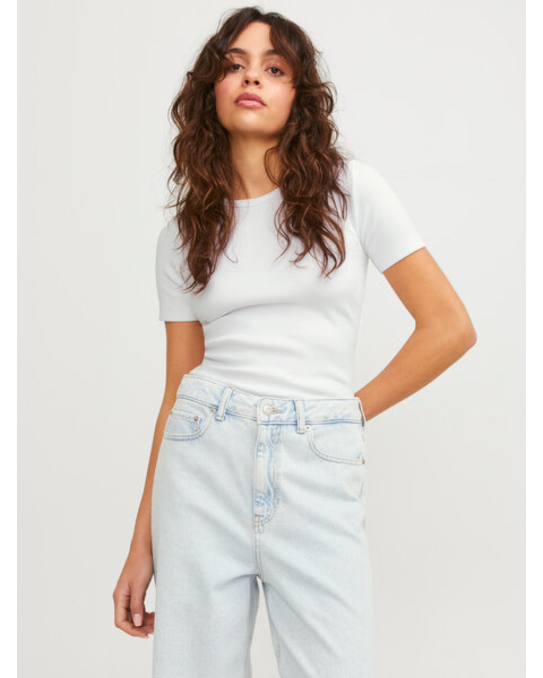 En person står afslappet iført en hvid T-shirt og lyse denim jeans. Konteksten er en simpel, lys baggrund. Ingen tekst er til stede.