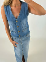 En person iført en ærmeløs denimvest med knapper og en matchende denimnederdel med slids. Personen har et armbånd og en halskæde på, mod en lys baggrund.