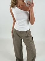 En person iført en hvid, asymmetrisk top og beige bukser tager et spejlbillede med en smartphone i et neutralt indendørs miljø.