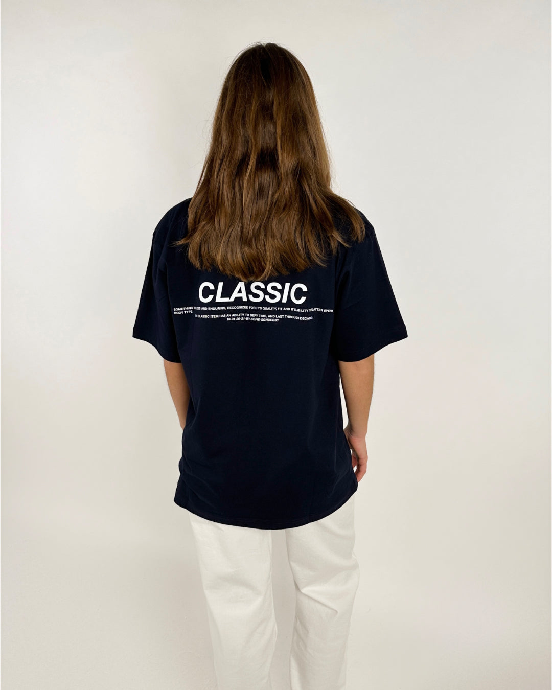 Person i sort T-shirt står med ryggen til, iført hvide bukser. T-shirten har teksten: "CLASSIC" efterfulgt af mindre tekst. Baggrunden er lys og enkel.