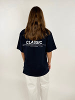 Person i sort T-shirt står med ryggen til, iført hvide bukser. T-shirten har teksten: "CLASSIC" efterfulgt af mindre tekst. Baggrunden er lys og enkel.
