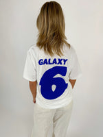 En person ses bagfra i en hvid T-shirt med blå tekst "GALAXY 6" i et lyst rum. Personen står stille med hænderne ved siden.