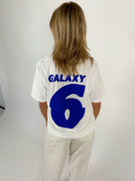 En person med skulderlangt hår står med ryggen til, iført en hvid t-shirt med blå tekst "GALAXY 6" i en lys baggrund.