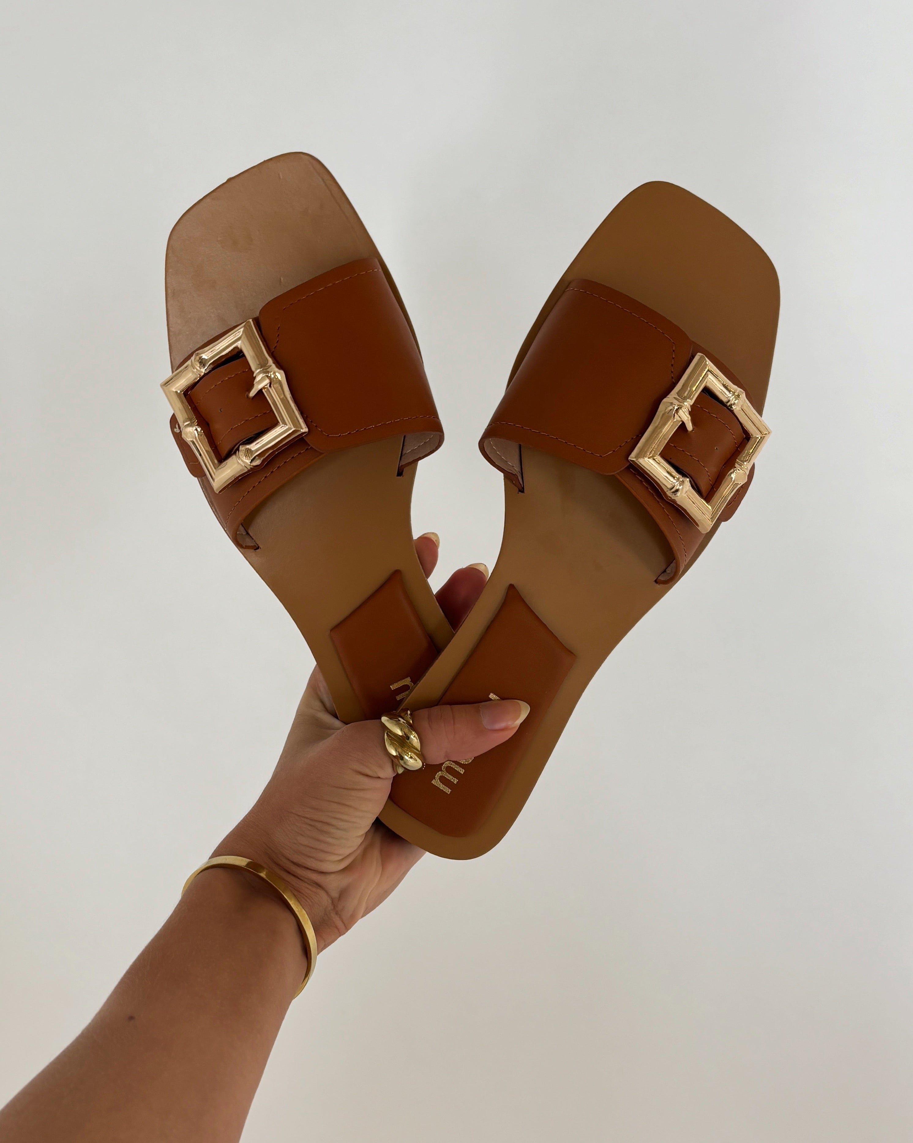 Brune sandaler med firkantede guldfarvede spænder holdes op af en hånd. Baggrunden er ensfarvet lys. Der er ingen synlig tekst.