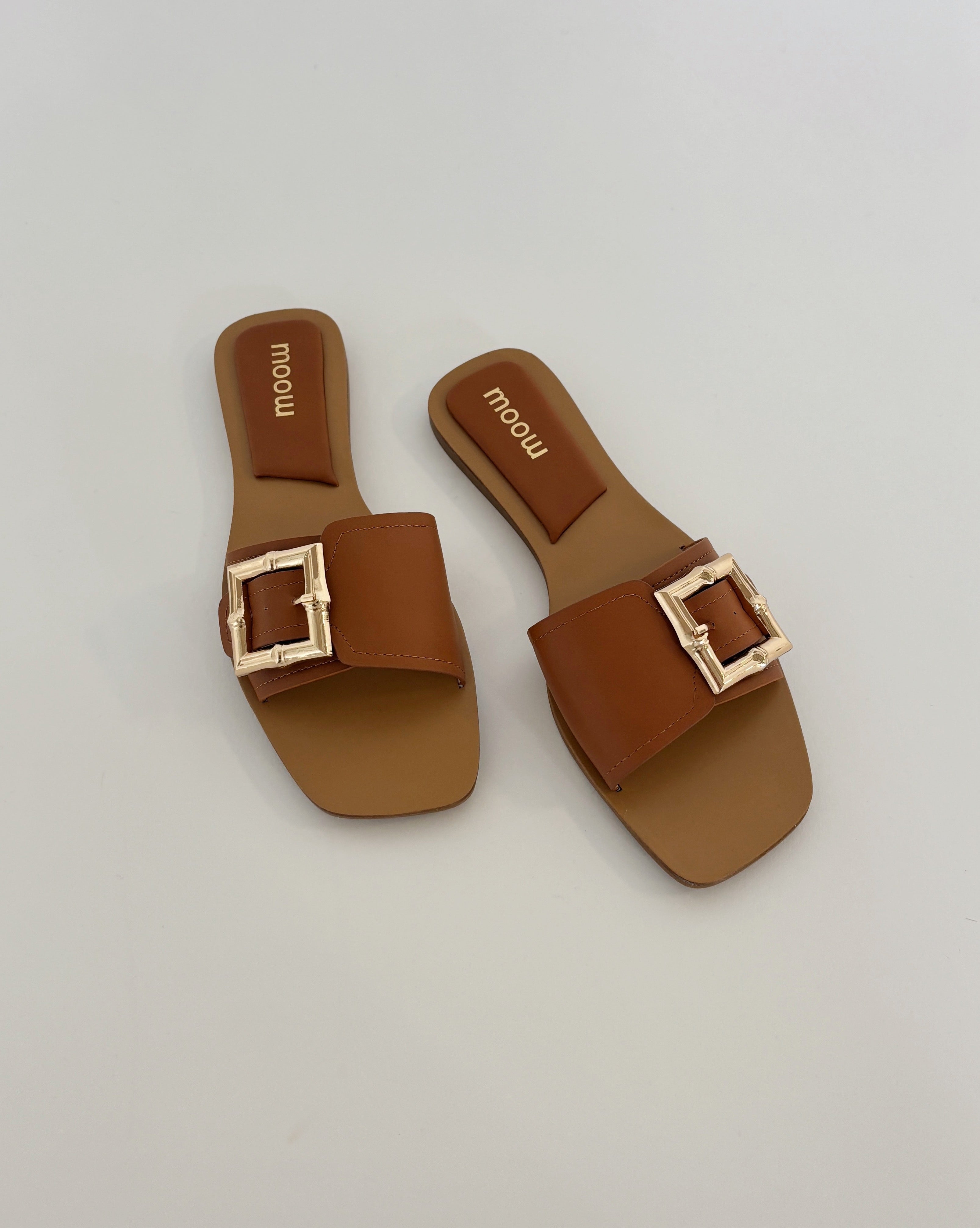 To brune sandaler med store, firkantede spænder ligger på en flad, hvid overflade. Mærket "moow" er trykt indeni sålerne.