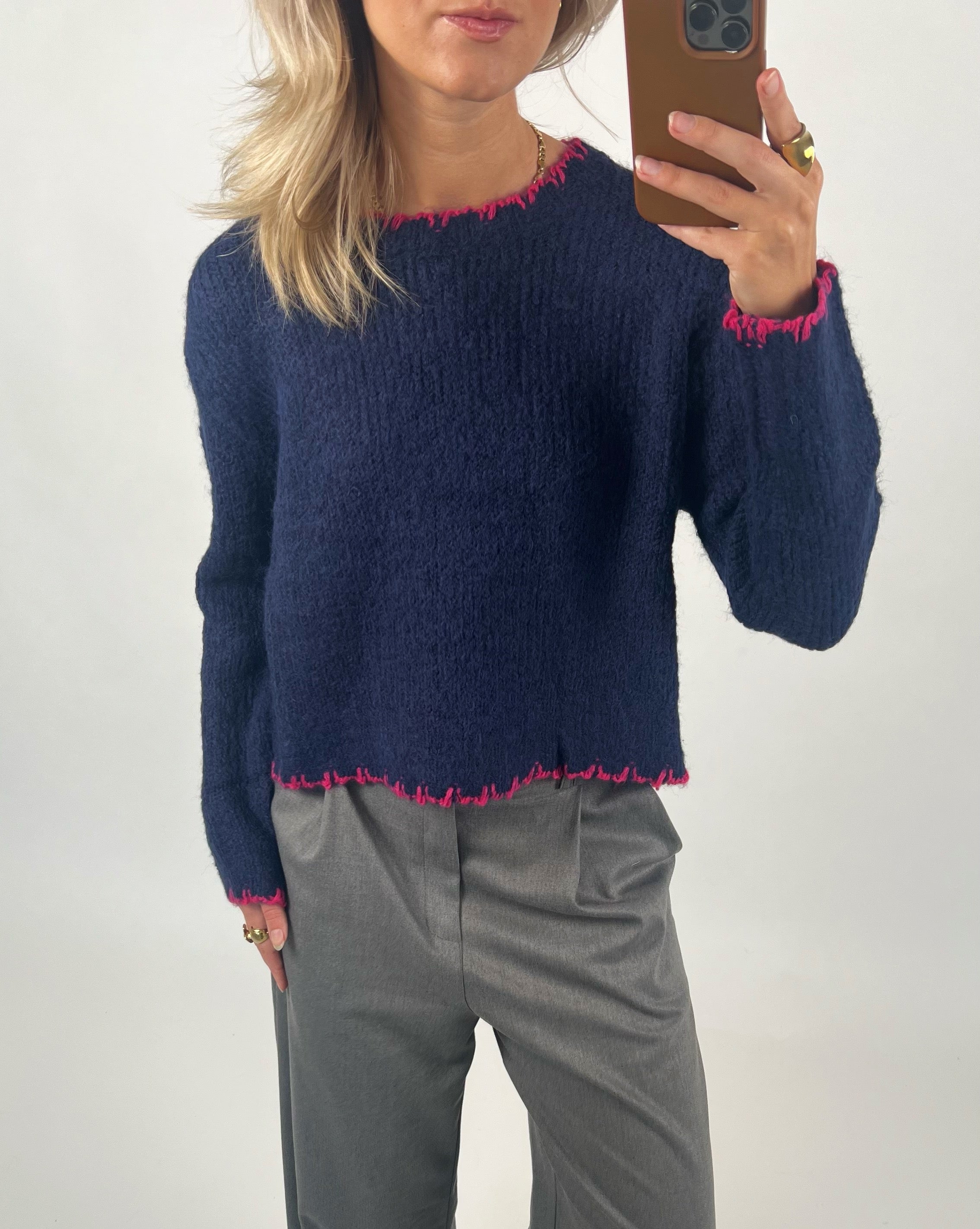 En person iført en mørkeblå sweater med pink kant detaljer holder en smartphone i hånden. Baggrunden er neutral, og personen bærer grå bukser og har lyst hår.