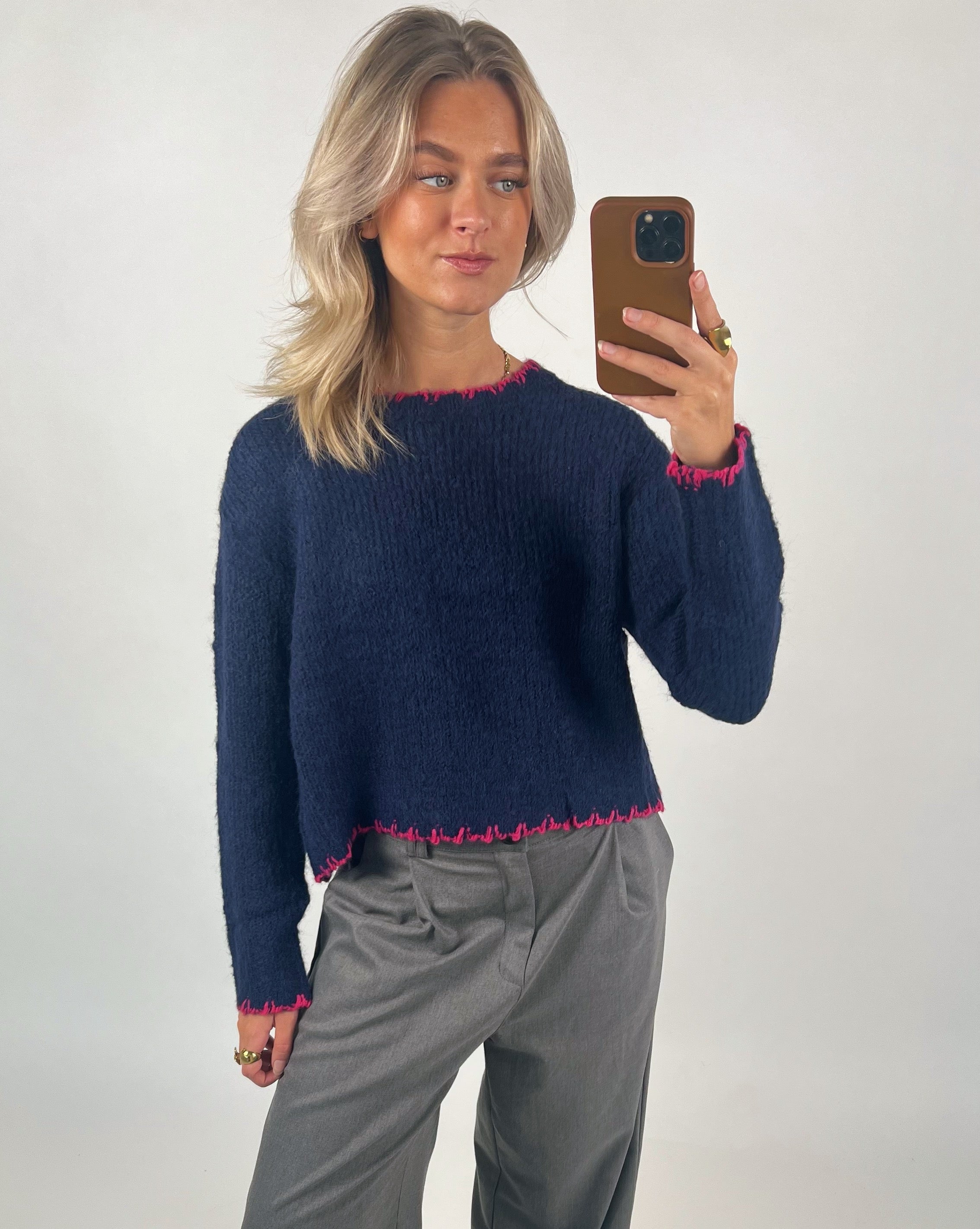 En person tager en selfie iført en mørkeblå trøje med lyserøde kanter og grå bukser. Personen står foran en lys baggrund og holder en brun mobiltelefon.