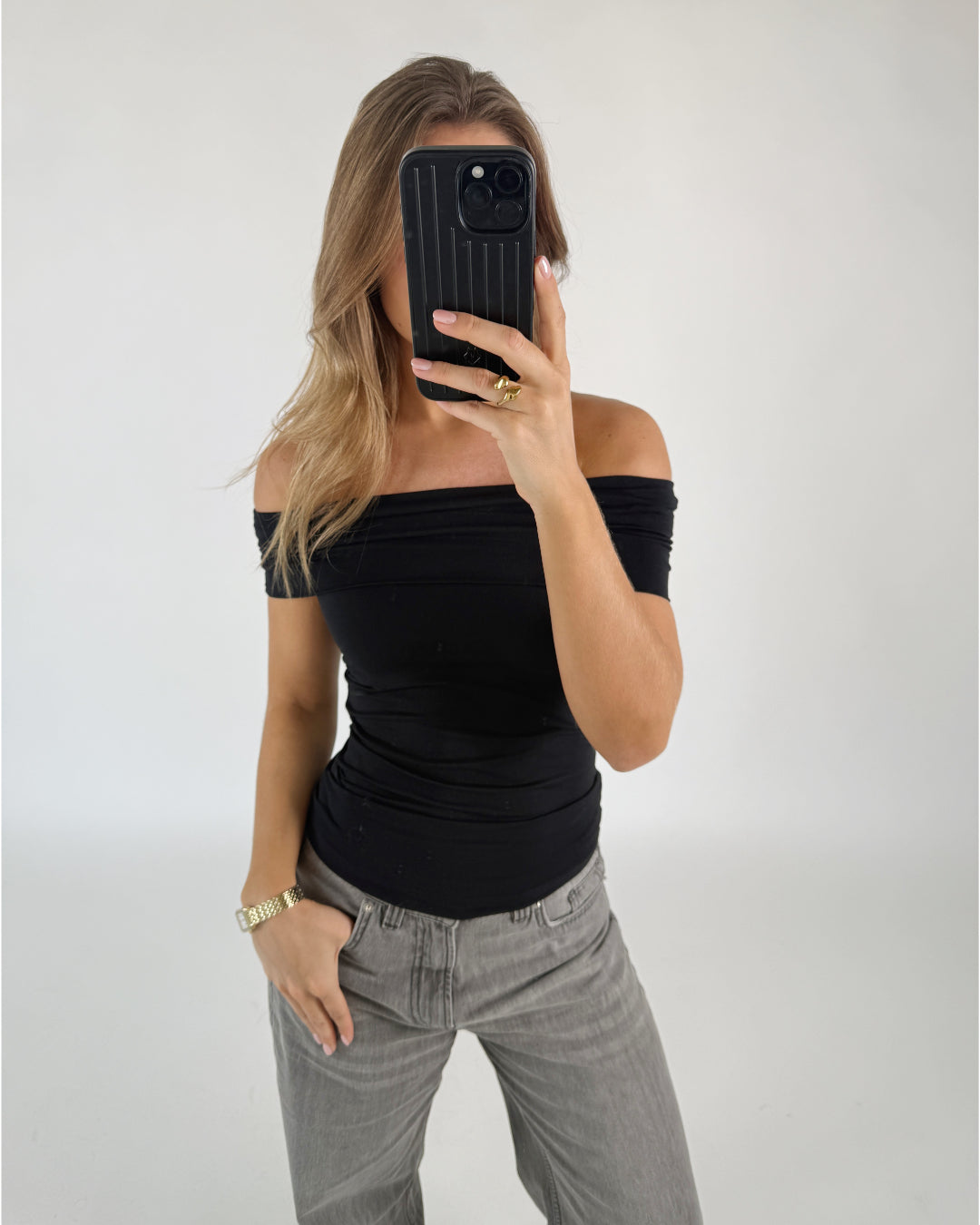 Person tager et spejlbillede iført en sort off-shoulder-top og grå bukser, i et enkelt rum. Personen holder en sort telefon op til ansigtet.