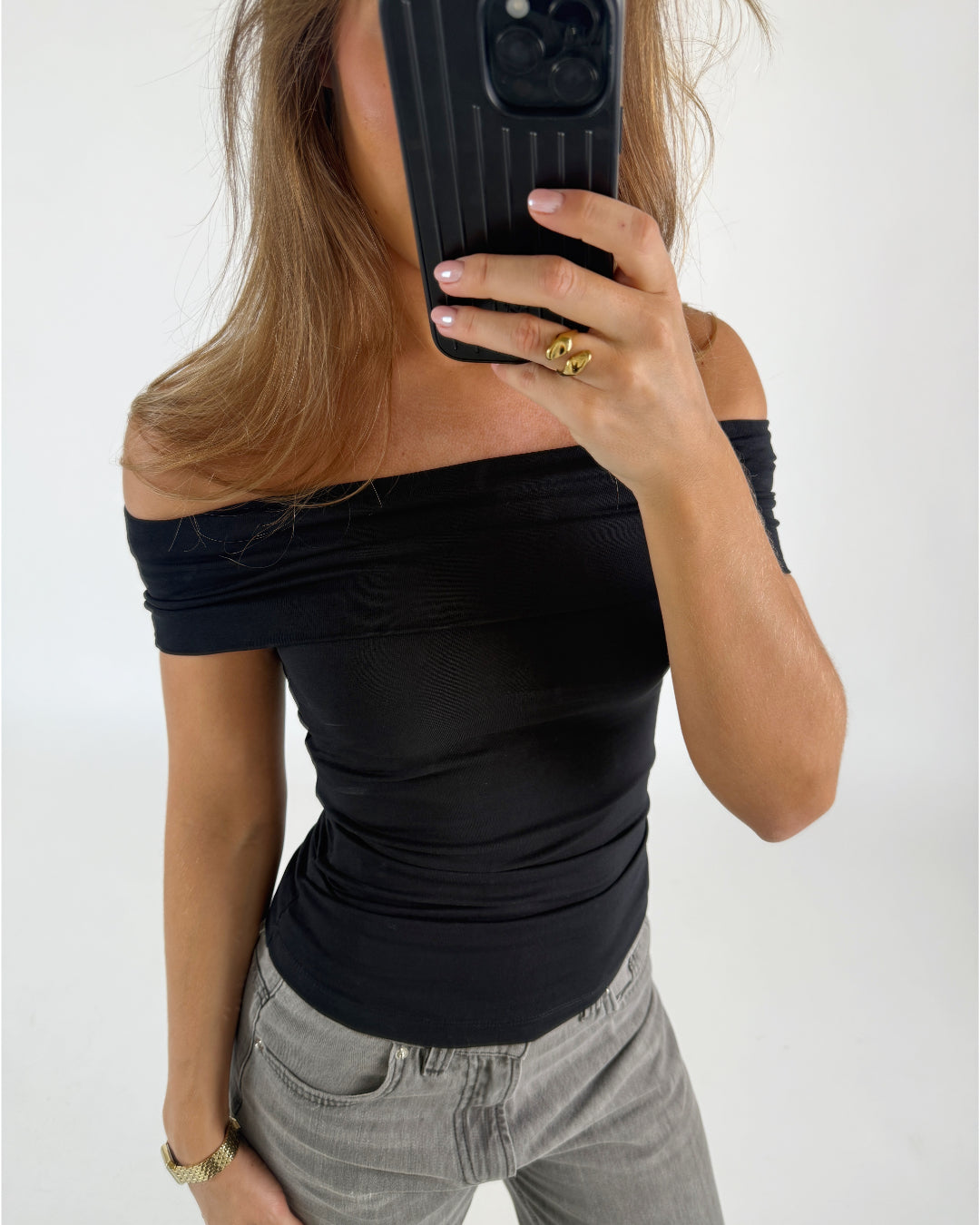Person tager et spejlbillede iført sort off-shoulder top og grå jeans. De holder en telefon foran ansigtet i en lys, minimalistisk baggrund.