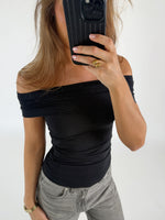 Person tager et spejlbillede iført sort off-shoulder top og grå jeans. De holder en telefon foran ansigtet i en lys, minimalistisk baggrund.