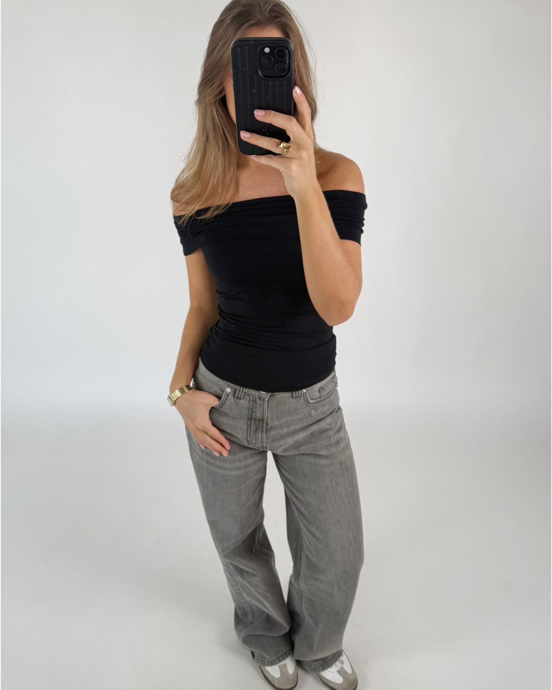 En person tager et spejlbillede, iført en sort, skulderfri top og grå jeans, mens de står i et minimalistisk, lyst rum med en neutral væg.