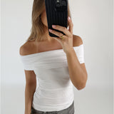 En person med en hvid off-shoulder top holder en sort smartphone foran ansigtet. Personen står i et lyst rum, iført grå jeans og en guld ring.