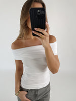 En person med en hvid off-shoulder top holder en sort smartphone foran ansigtet. Personen står i et lyst rum, iført grå jeans og en guld ring.