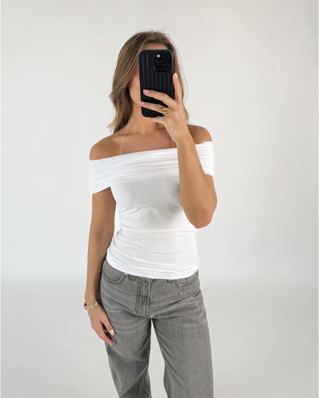 Person iført en hvid off-shoulder top og grå bukser tager et spejlfoto med en sort telefon. Baggrunden er en simpel, hvid væg.
