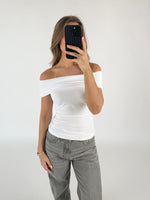 Person iført en hvid off-shoulder top og grå bukser tager et spejlfoto med en sort telefon. Baggrunden er en simpel, hvid væg.