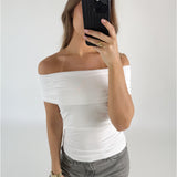 Person i hvid top tager et billede med en sort telefon, iført grå jeans og smykker, står i et minimalistisk rum.