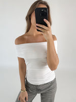 Person i hvid top tager et billede med en sort telefon, iført grå jeans og smykker, står i et minimalistisk rum.