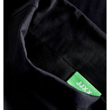 Sort stof med grøn etiket viser mærketeksten "JAXX" i hvid. Etiketten er delvist synlig, omgivet af mørkt, foldet tekstil.