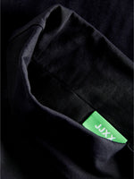 Sort stof med grøn etiket viser mærketeksten "JAXX" i hvid. Etiketten er delvist synlig, omgivet af mørkt, foldet tekstil.