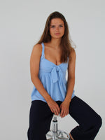 En person sidder på en barstol og kigger fremad. Personen bærer en blå top og mørke bukser i et enkelt studie med en hvid baggrund.