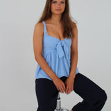 En kvinde sidder afslappet på en barstol. Hun bærer en lyseblå top og mørke bukser i et neutralt rum med hvid baggrund.