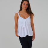 En kvinde iført en hvid top med en sløjfe foran og sorte bukser står foran en neutral hvid baggrund. Hun har hånden i bukselommen.