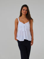 Kvinde iført hvid top står roligt, med hænderne langs siden, på en neutral, hvid baggrund. Hun har løst, langt hår og sorte bukser.