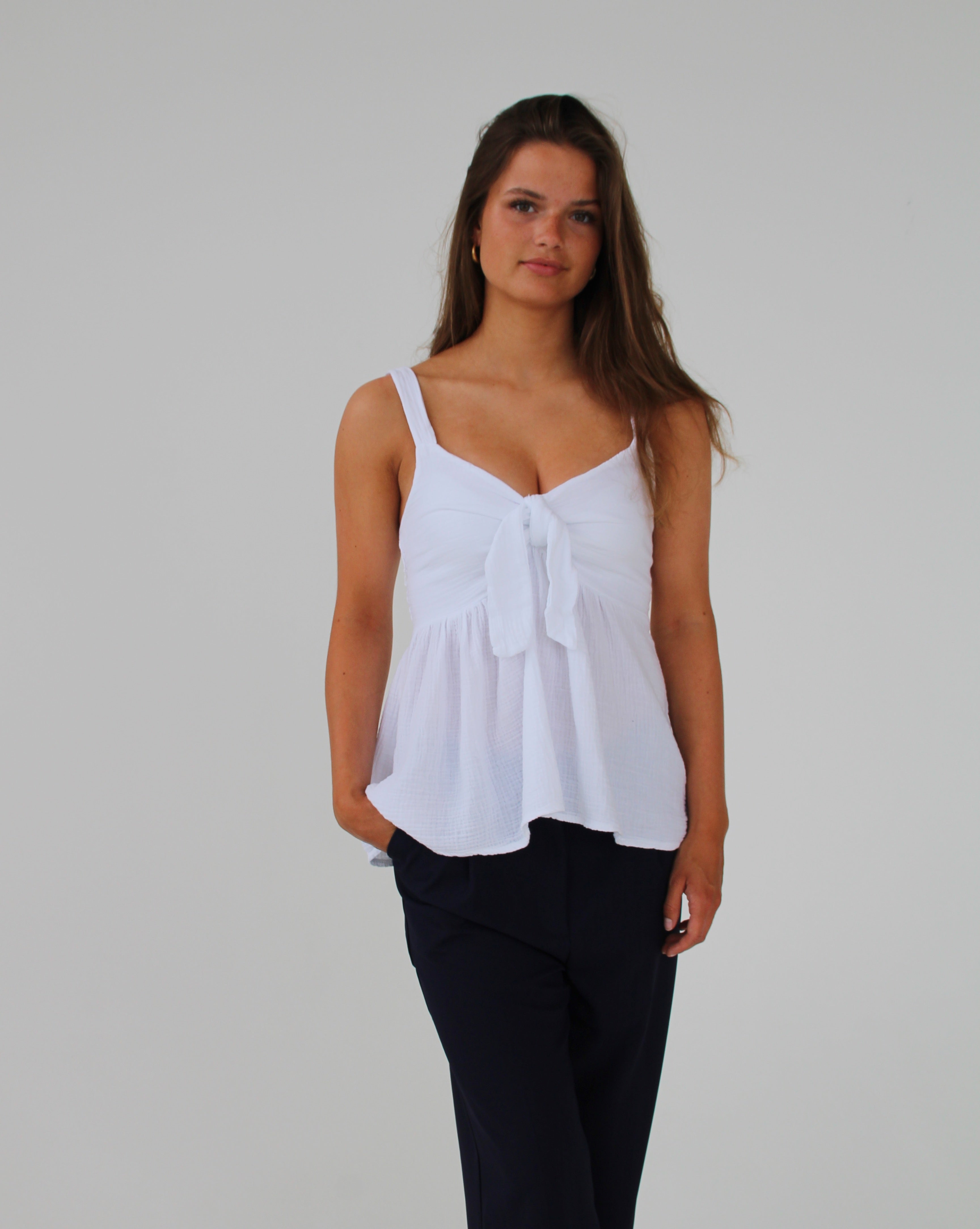 Kvinde iført hvid top står roligt, med hænderne langs siden, på en neutral, hvid baggrund. Hun har løst, langt hår og sorte bukser.