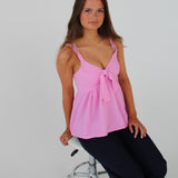 En kvinde sidder på en hvid, polstret taburet iført en lyserød top og mørke bukser. Baggrunden er ensfarvet hvid, hvilket fremhæver hendes positur.