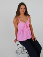 En kvinde sidder på en hvid, polstret taburet iført en lyserød top og mørke bukser. Baggrunden er ensfarvet hvid, hvilket fremhæver hendes positur.